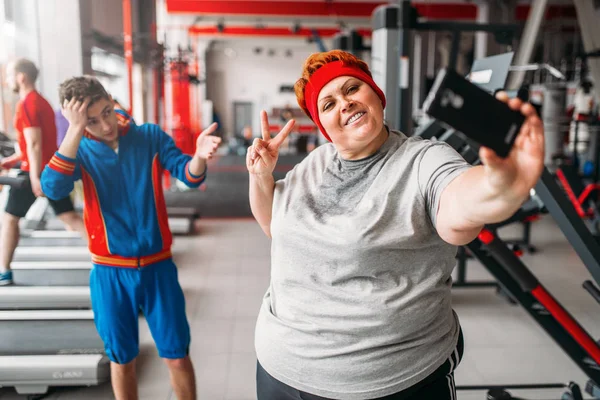 fat sweaty woman taking selfie with skinny male instructor in gym