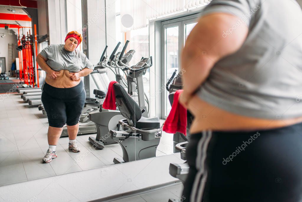 overweight sweaty woman looking at her body against mirror in gym
