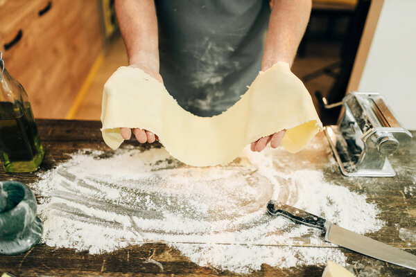 man in apron working with dough, pasta machine on wooden kitchen table