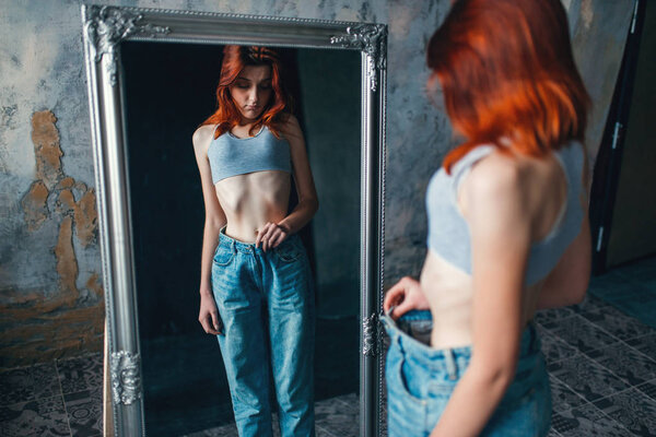 thin woman trying on big size jeans against mirror, weight loss, anorexia