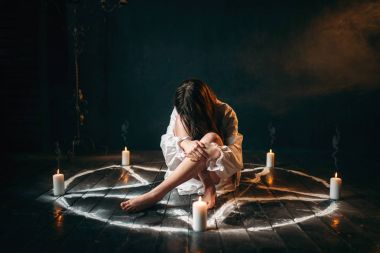 young woman in white shirt sitting in center of pentagram circle with candles, ritual of black magic, black wooden floor