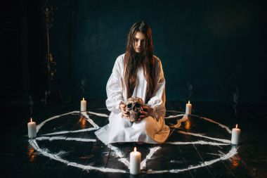 young woman in white shirt holding human skull in pentagram circle 