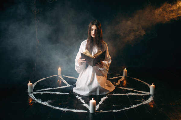 young woman in white shirt sitting in the center of pentagram circle with candles and reading a spell, dark magic ritual, occultism