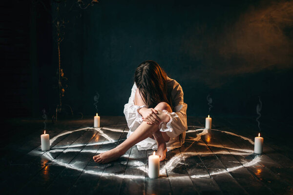 молодая женщина в белой рубашке, сидящая в центре пентаграммного круга со свечами, ритуал черной магии, черный деревянный пол
