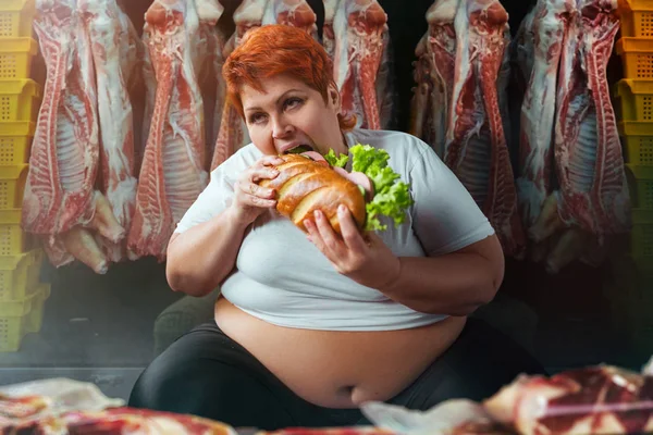fat woman eating big burger against meat carcasses, overweight concept
