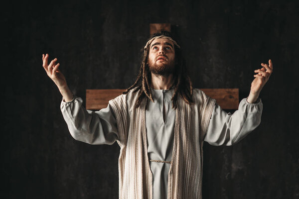Jesus Christ in white robe emotionally prays with his hands up, dark background. Son of God, strong faith in God