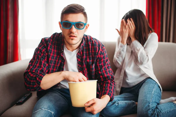 Couple sitting on the couch, man in 3D glasses with popcorn in hands watch movie, frightened woman closes her face with hands