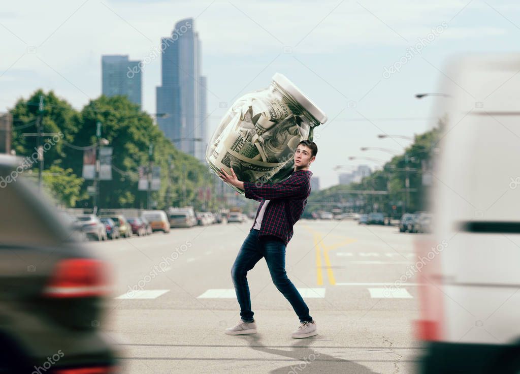 Money saving concept, young man trying to carry a big glass jar full of dollars. Cash economy addiction, home bank
