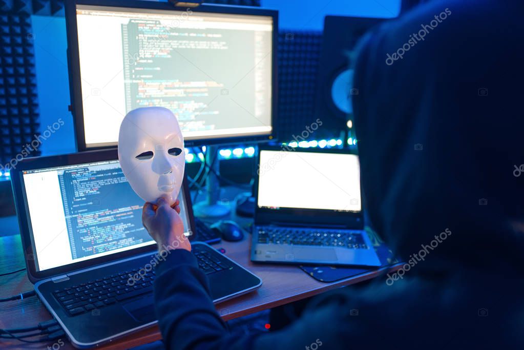 Hacker in hood holds mask in hand and at his workplace with laptop and PC, password or account hacking. Internet spy, crime lifestyle, risk job, network criminal