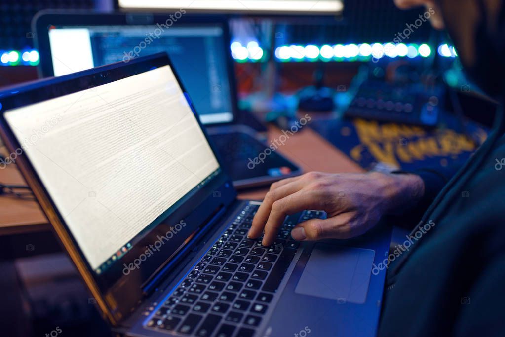 Hacker in hood shows thumbs up at his workplace with laptop and PC, password or account hacking. Internet spy, crime lifestyle, risk job, network criminal