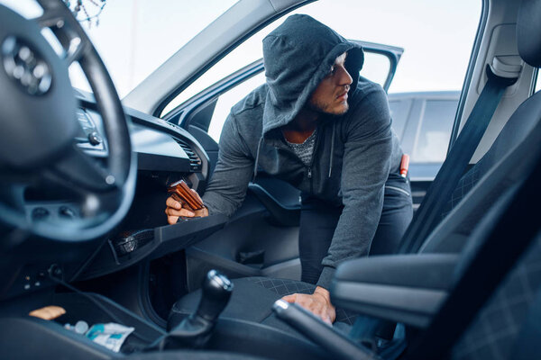 Car robber takes the wallet from the glove compartment, criminal lifestyle, stealing. Hooded male bandit opening vehicle on parking. Auto robbery