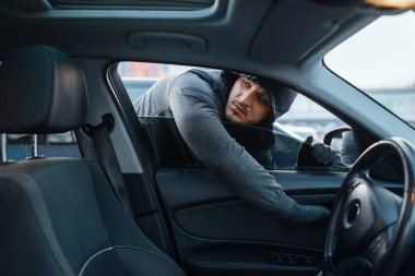 Araba hırsızı kapıyı açıyor, risk alıyor, hırsızlık yapıyor. Kapüşonlu erkek haydut otoparkta araca biniyor. Otomobil soygunu, otomobil suçu