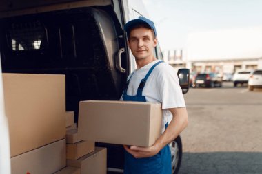 Arabada karton kutusuyla gülümseyen teslimatçı, teslimat servisi. Üniformalı bir adam karton bir paket taşıyor, erkek teslimatı, kurye işi.