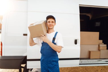 Deliveryman in uniform holds carton box at the car, delivery service. Man holding cardboard package, male deliver, courier or shipping job clipart