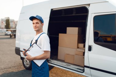 Paket ve defter taşıyan üniformalı teslimatçı, arabada karton kutular, teslimat servisi. Karton paketlerin başında duran bir adam araç, erkek teslimatı, kurye ya da nakliye işi