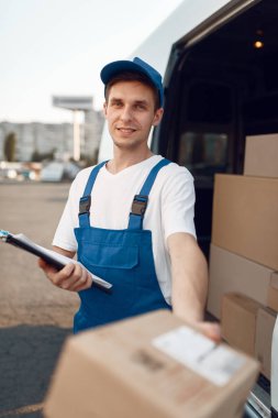 Üniformalı teslimatçı paketini, karton kutusunu, teslimat servisini verir. Karton paketlerin başında duran bir adam araç, erkek teslimatı, kurye ya da nakliye işi