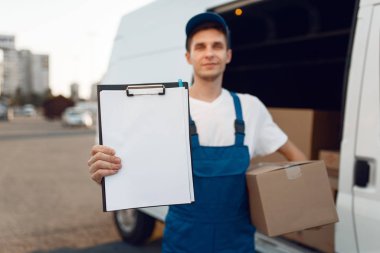 Paket ve defter taşıyan üniformalı teslimatçı, arabada karton kutular, teslimat servisi. Karton paketlerin başında duran bir adam araç, erkek teslimatı, kurye ya da nakliye işi