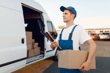 Üniformalı teslimatçı paketini, karton kutusunu, teslimat servisini verir. Karton paketlerin başında duran bir adam araç, erkek teslimatı, kurye ya da nakliye işi