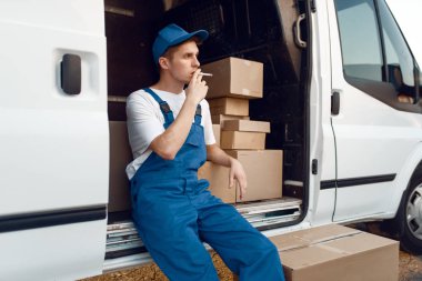 Mola sırasında üniformalı teslimatçı arabada sigara içiyor, paketli ve karton kutulu otomobil, teslimat servisi. Karton paketlerin başında duran bir adam araç, erkek teslimatı, kurye ya da nakliye işi