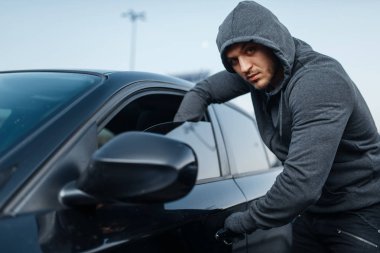Araba hırsızlığı, kapı kırmak, suç işlemek, hırsızlık, hırsızlık. Kapüşonlu erkek soyguncu park yerinde araç açıyor. Araba soygunu, otomobil suçu, vandalizm