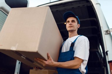 Karton kutu taşıyan üniformalı bir yükleyici, teslimat servisi. Karton paketlerin başında duran bir adam araç, erkek teslimatı, kurye ya da nakliye işi