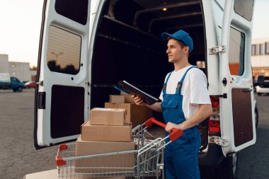 Üniformalı teslimatçı, paketli kutularla dolu el arabasını arabada tutuyor, teslimat servisi. Karton paketlerin başında duran bir adam araç, erkek teslimatı, kurye ya da nakliye işi