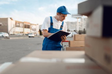 Üniformalı teslimatçı. Paketleri kontrol et, teslimat servisi. Karton paketlerin başında duran bir adam araç, erkek teslimatı, kurye ya da nakliye işi