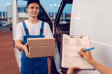 Üniformalı kurye arabadaki kadın alıcıya bir paket veriyor, teslimat servisi. Aracın yanında karton paket taşıyan adam, erkek teslimatı ve kadın, kurye ya da nakliye işi