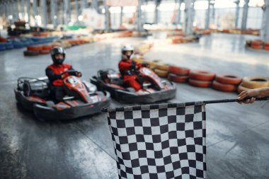 Başlangıç çizgisinde iki kart yarışçısı, kareli bayrak, ön görüş, kapalı alanda karting otomobil sporu. Lastik bariyeriyle yakın go-cart pistinde hız yarışı. Hızlı araç yarışması, yüksek adrenalin eğlencesi.