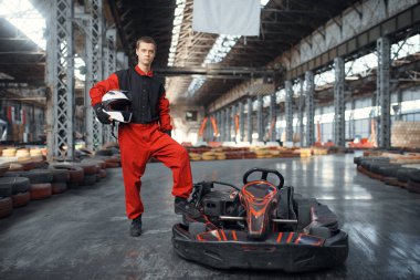 Miğferli yarışçı go kart arabasının yanında poz veriyor, kapalı mekanda araba sporu yapıyor. Lastik engelli go-kart pistinde hız yarışı. Hızlı araç yarışması, yüksek adrenalin eğlencesi.