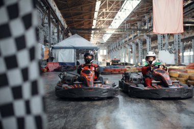 Başlangıç çizgisinde iki kart yarışçısı, kareli bayrak, ön görüş, kapalı alanda karting otomobil sporu. Lastik bariyeriyle yakın go-cart pistinde hız yarışı. Hızlı araç yarışması, yüksek adrenalin eğlencesi.
