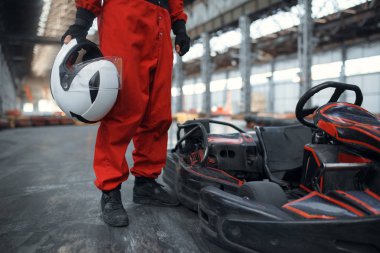 Miğferli yarışçı go kart arabasının yanında poz veriyor, kapalı mekanda araba sporu yapıyor. Lastik engelli go-kart pistinde hız yarışı. Hızlı araç yarışması, yüksek adrenalin eğlencesi.
