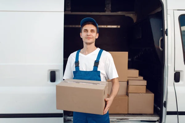 Deliveryman with carton box at the car, delivery service. Man in uniform holding cardboard package, male deliver, courier job