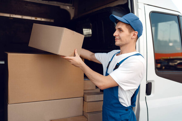 Smiling deliveryman with carton box at the car, delivery service. Man in uniform holding cardboard package, male deliver, courier job