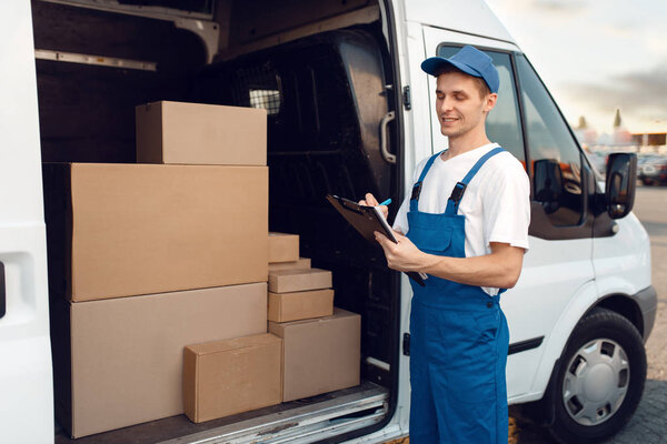 Deliveryman in uniform, carton boxes in the car, delivery service. Man standing at cardboard packages in vehicle, male deliver, courier or shipping job