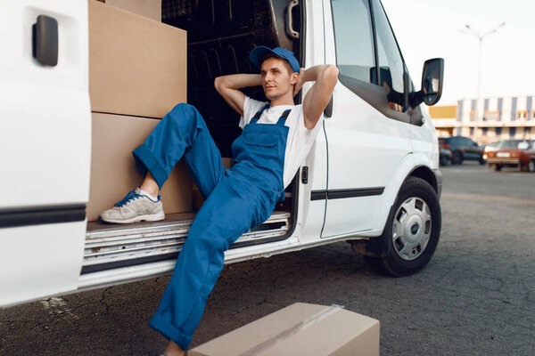 Deliveryman in uniform relaxing in the car during a break, auto with parcels and carton boxes, delivery service. Man poses at cardboard packages in vehicle, male deliver, courier or shipping job