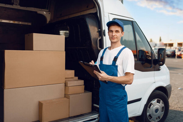 Deliveryman in uniform, carton boxes in the car, delivery service. Man standing at cardboard packages in vehicle, male deliver, courier or shipping job