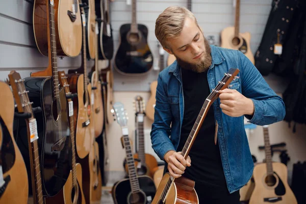 音楽店でアコースティックギターを選ぶ髭の若い男 楽器店の依頼 男性音楽家 — ストック写真