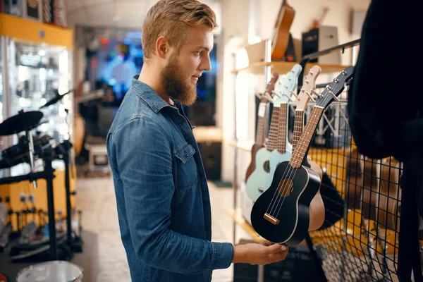 音楽店でウクレレギターを選ぶ髭の若い男 楽器店での品揃え 男性音楽家購入機器 — ストック写真