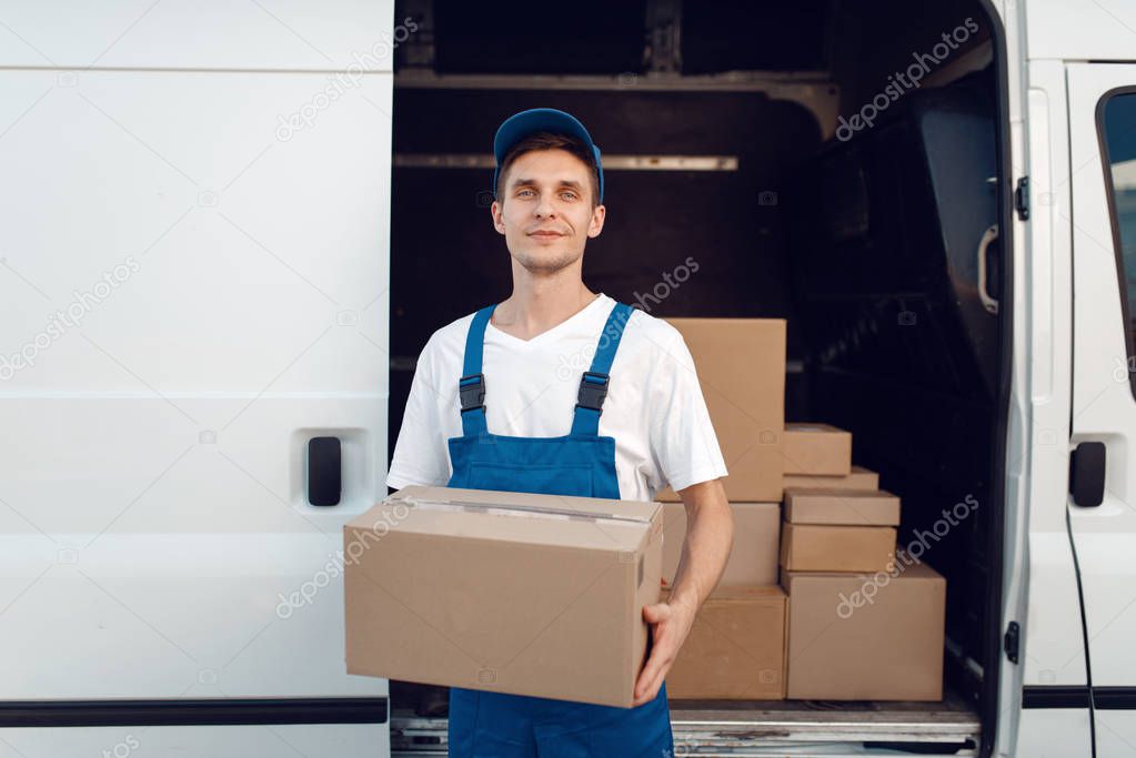 Deliveryman with carton box at the car, delivery service. Man in uniform holding cardboard package, male deliver, courier job