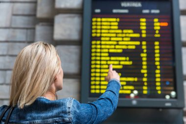 Female tourist looks on timetable on railway station, travel in Europe. Transportation by european railroads, passenger at the information display, comfortable tourism and travelling clipart