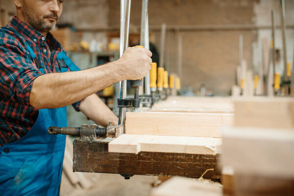 Плотник в форме зажимает доску в тисках, деревообработке, деревообрабатывающей промышленности, плотницкой промышленности. Деревообработка на мебельной фабрике, производство продукции из натуральных материалов
