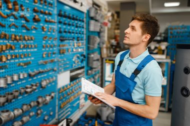 Tesisatçı dükkanında kapakçıkları ve boru adaptörleri olan üniformalı bir tesisatçı. Defterli bir adam dükkandan hijyen mühendisliği satın alıyor.