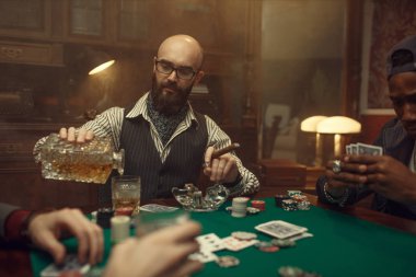 Sakallı poker oyuncusu elinde puroyla viski ve kumarhane dolduruyor. Şans oyunları bağımlılığı. Kumarhanede adam boş vakit geçirir. Oyun masasında yeşil kumaşlı kartlar ve cipsler.