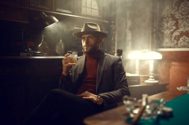 Poker oyuncusu takım elbiseli ve şapkalı kumarhane barında viski içiyor, bağımlılık riski var. Şans oyunları işte. Kumarhanede boş vakitler, yeşil kumaşlı kumar masası.