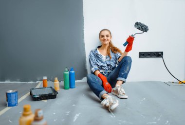 İşten sonra yerde boya silindirli kadın boyacı oturuyor. Ev onarımı, mutlu bir kadın apartman tamiri yapıyor.