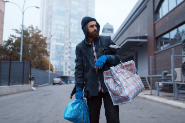 Bearded tramp man with bag on city street. Poverty is a social problem, homelessness and loneliness, alcoholism and  drunk addiction, urban lonely