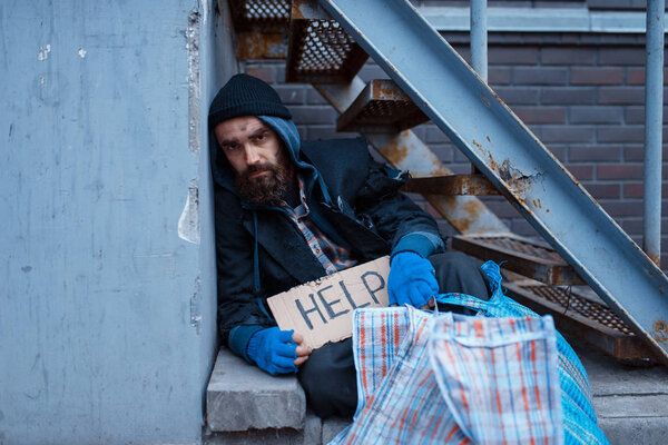 Bearded bum with help sign on city street. Poverty is a social problem, homelessness and loneliness, alcoholism and  drunk addiction, urban lonely