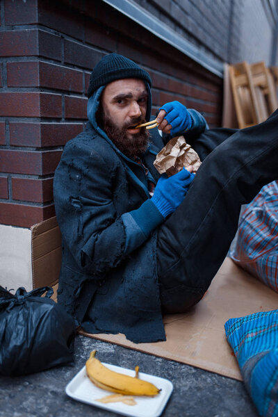 Bearded beggar eats burger on city street. Poverty is a social problem, homelessness and loneliness, alcoholism and drunk addiction
