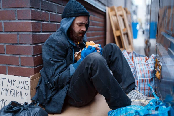 Bearded beggar eats burger on city street. Poverty is a social problem, homelessness and loneliness, alcoholism and drunk addiction